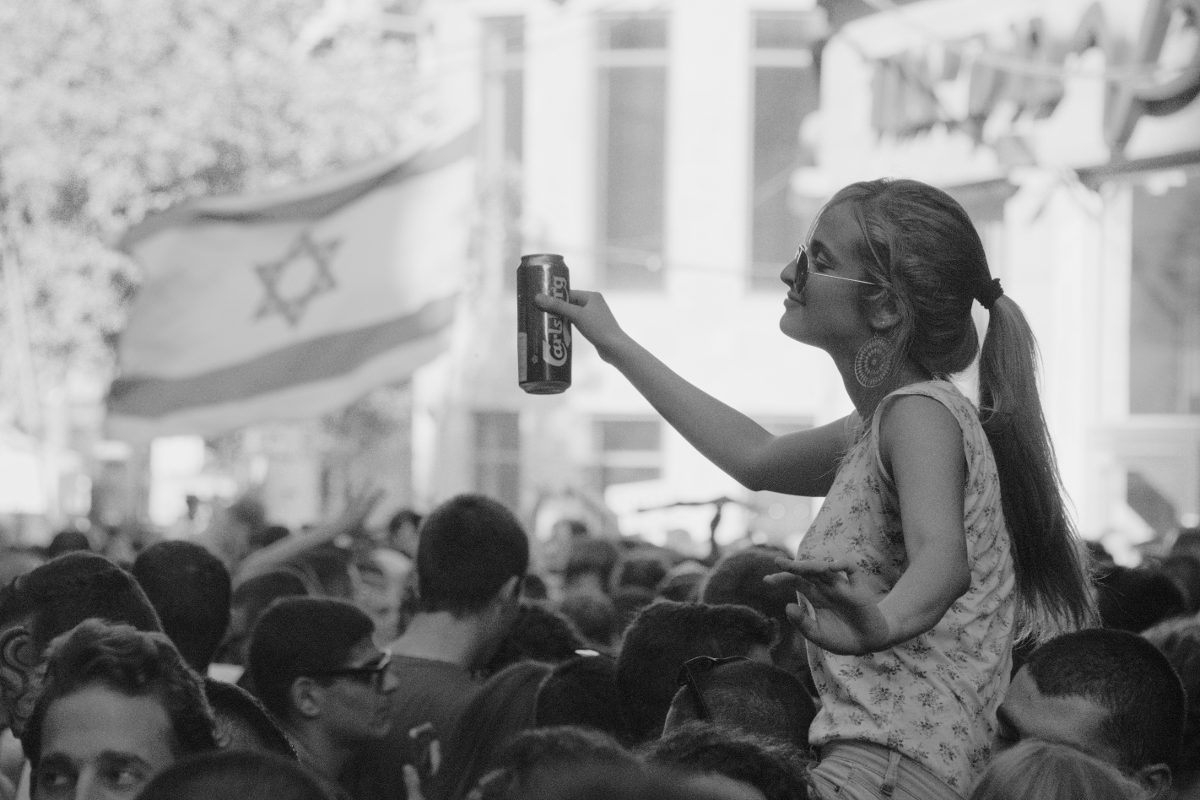 Une jeune femme dépasse d'une foule de fêtards, une bière à la main, le drapeau israélien à l'arrière-plan