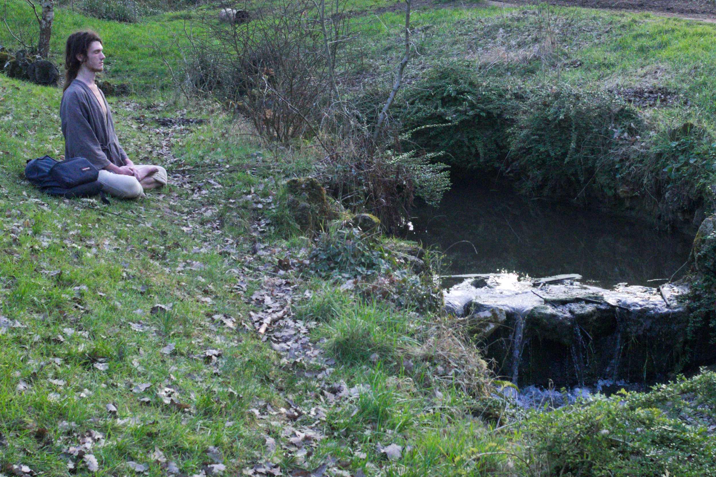 Un jeune homme médite en position du lotus, au bord d'un ruisseau, dans le bois de Vincennes.