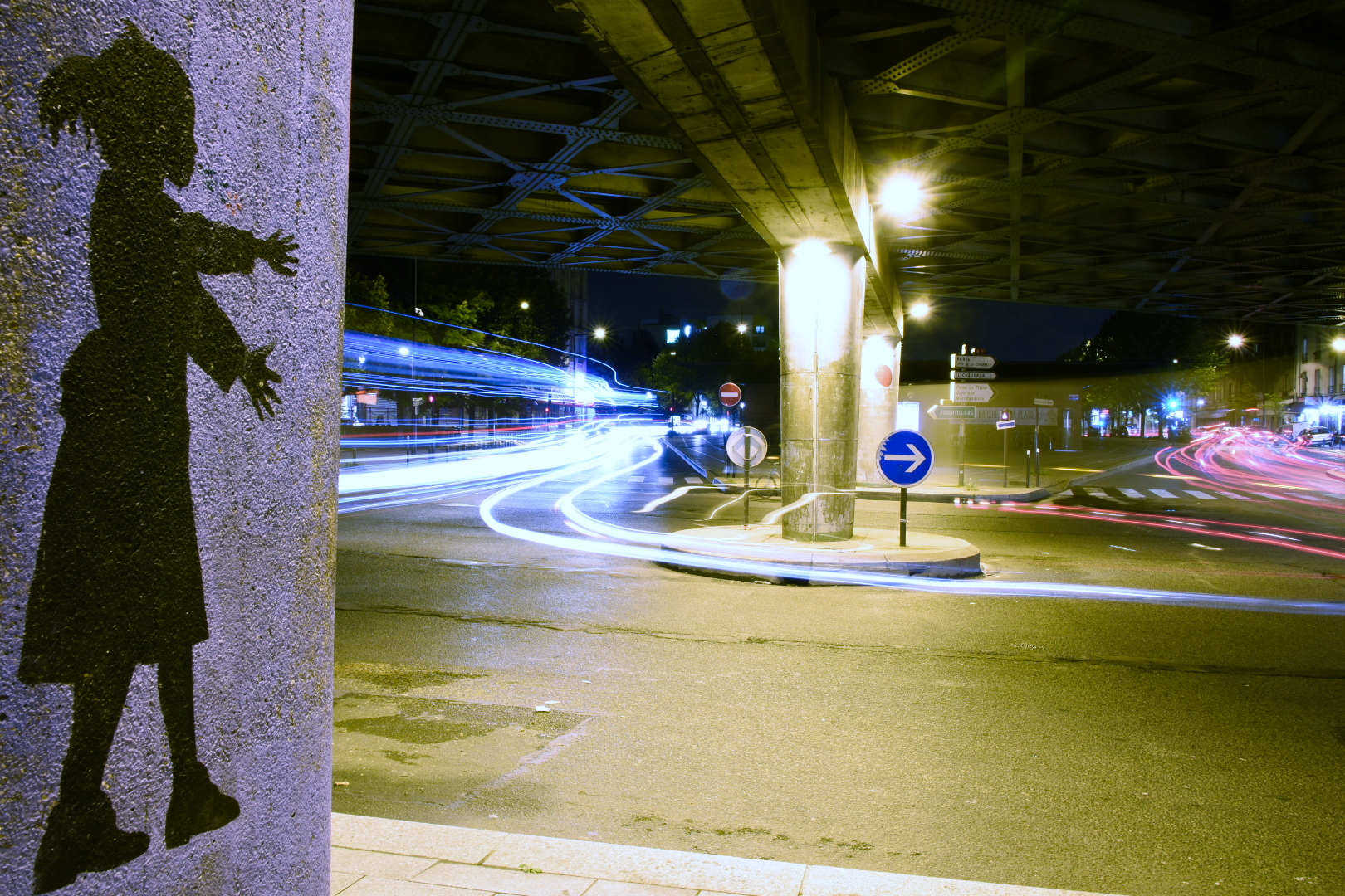 Circulation automobile de nuit, sous un pont autoroutier, avec un rond-point et un graffiti représentant une petite fille